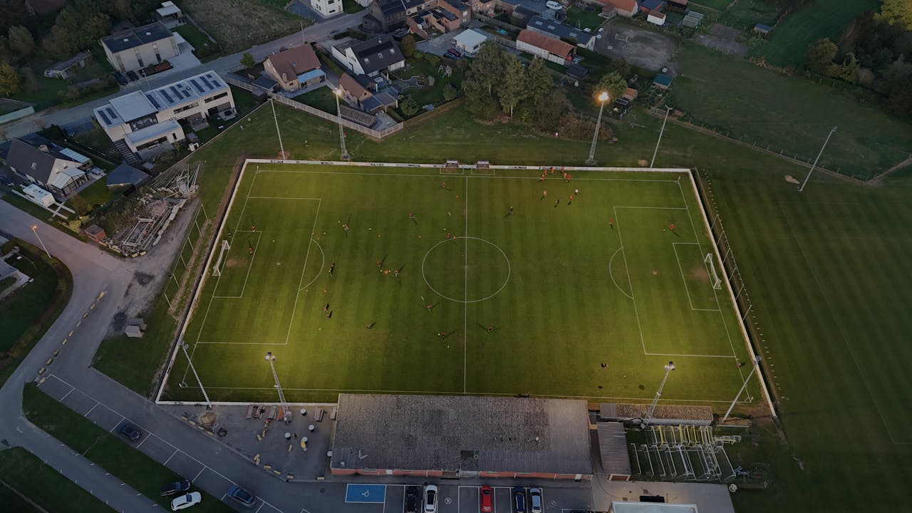 Aerial shot of a soccer field at dusk in België with players practicing under floodlights.